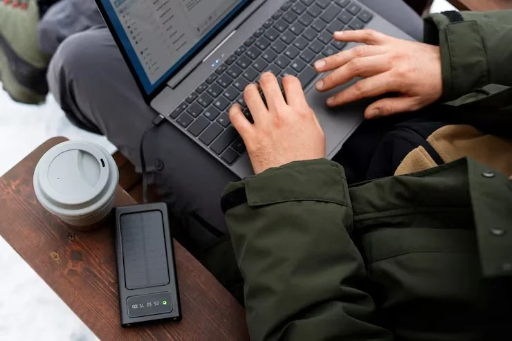 Persona tecleando en un portátil al aire libre, con una batería solar y un café